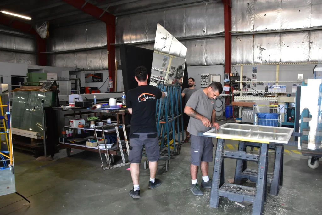 Dans l'atelier, un technicien changeant une vitre sur un cadre de fenêtre, deux techniciens transportant un plateau de miroir vers la machine à découpe