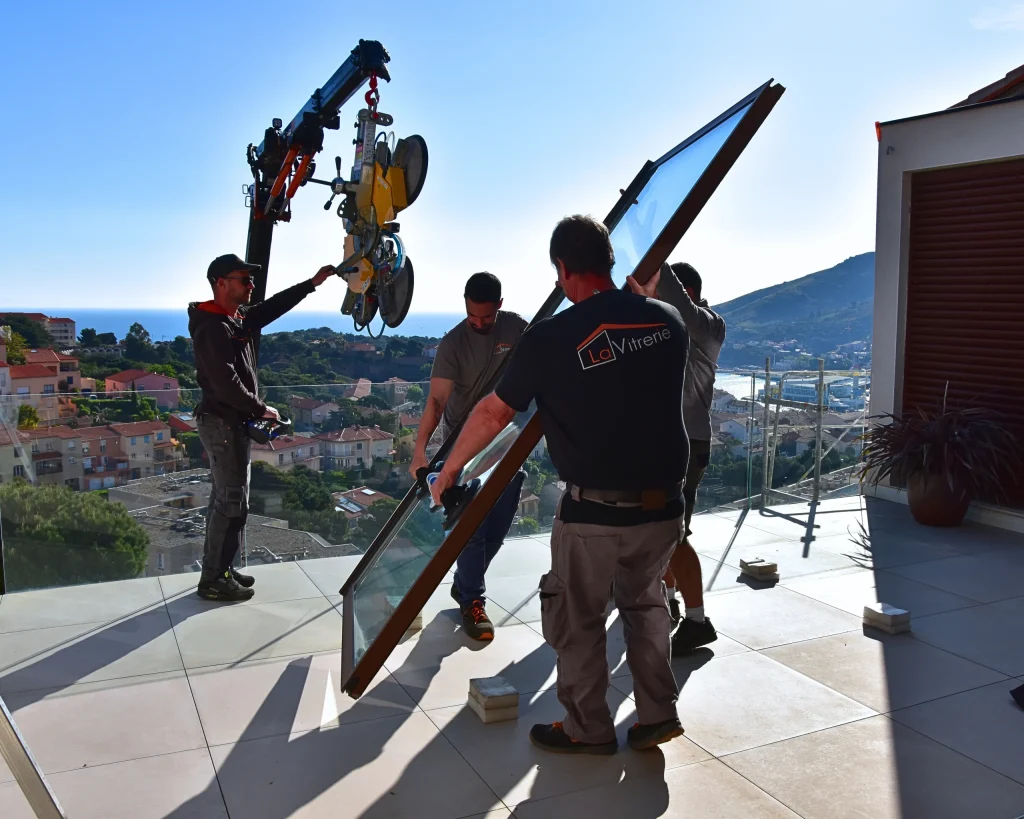 Prise de vue sur chantier, l'équipe de quatre techniciens aidé d'une grue afin de remplacer cette baie vitrée sur un balcon, vue sur Port Vendres et la Méditerranée
