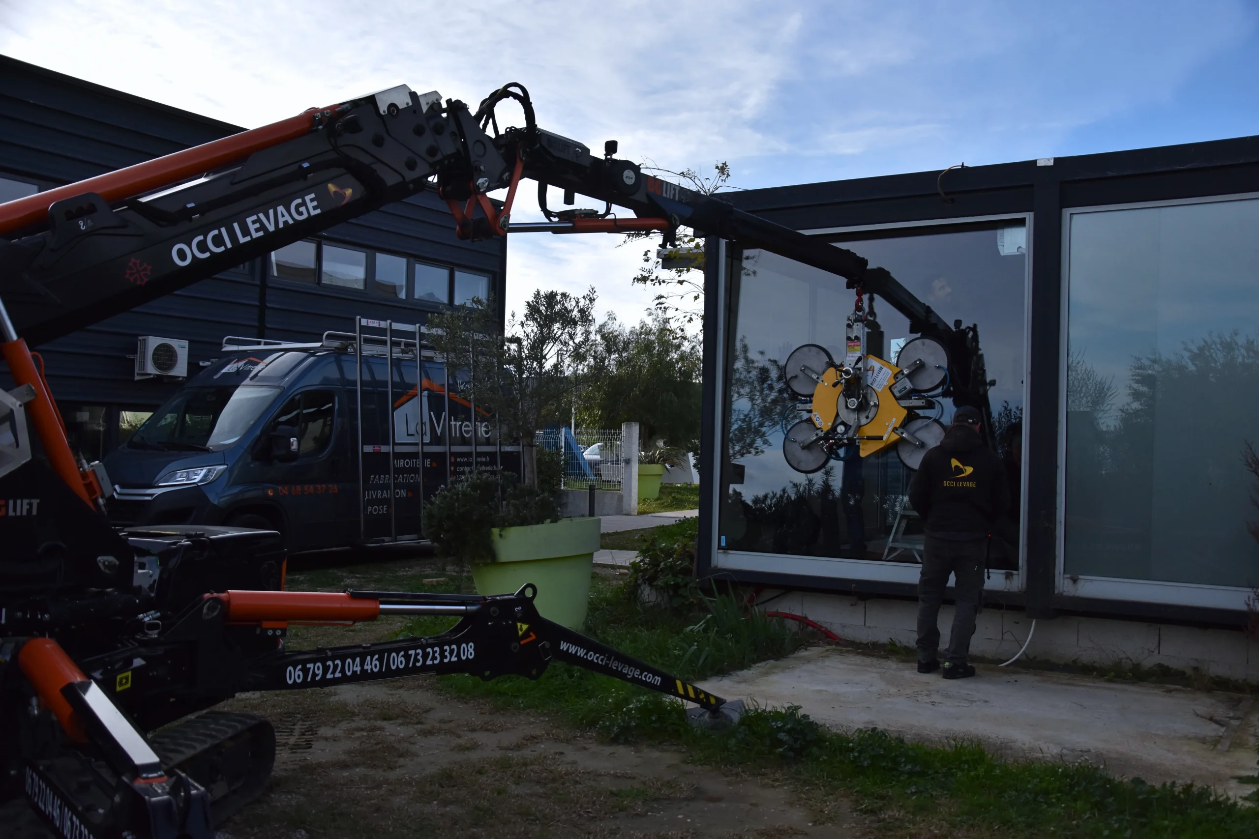 Sur chantier à l'Aéropyrénées, remplacement d'un grand double vitrage avec l'aide d'une grue, vue sur le bras de la grue en train d'enlever le verre avec le fourgon de La Vitrerie en arrière plan
